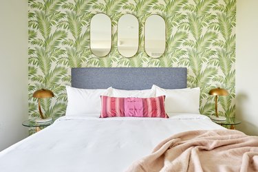 Eclectic bedroom with upholstered headboard, tropical print wallpaper, pink pillow, brass lamps.